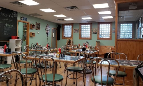 A cozy café interior with empty tables, green chairs, and wooden accents, featuring condiments on each table.