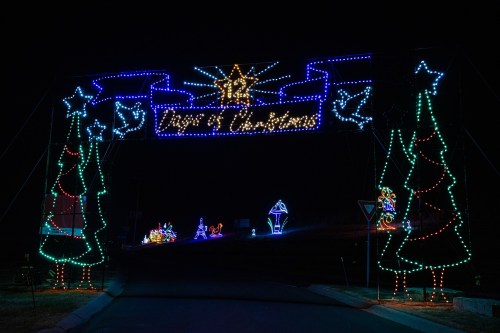 Colorful Christmas lights arch over a road, featuring trees, stars, and festive decorations in a nighttime setting.