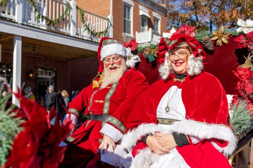 Santa Claus and Mrs. Claus smile while sitting together in a festive sleigh, surrounded by holiday decorations.
