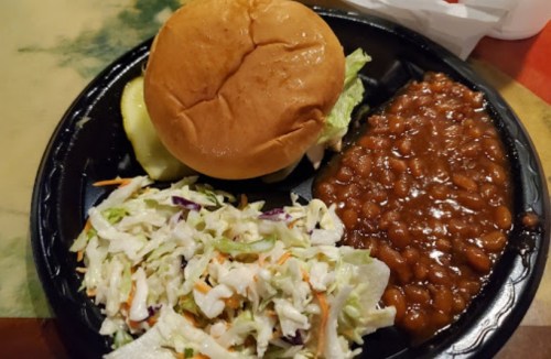 A plate with a hamburger, pickles, coleslaw, and baked beans on a black tray.