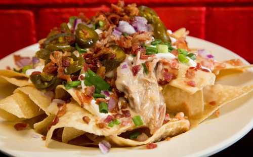 A plate of nachos topped with jalapeños, bacon, green onions, and sour cream against a red background.