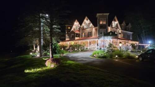 A beautifully lit house at night, surrounded by trees and flowers, with a welcoming porch and driveway.