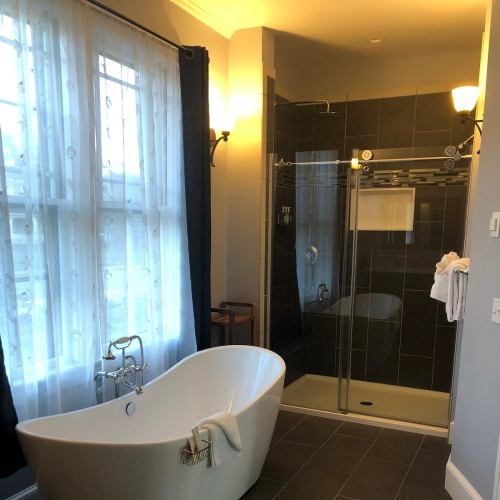 A modern bathroom featuring a freestanding bathtub, glass shower, and large window with blue curtains.
