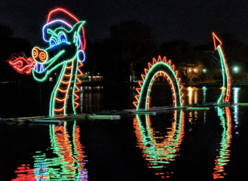 A colorful dragon-shaped light display with a Santa hat, reflecting in the water at night.