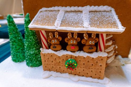 A festive gingerbread house with three reindeer figures in the window, surrounded by green trees and a wreath.