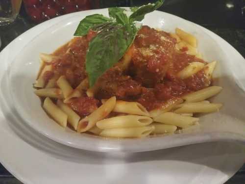 A plate of penne pasta topped with marinara sauce, meatballs, and a fresh basil leaf.