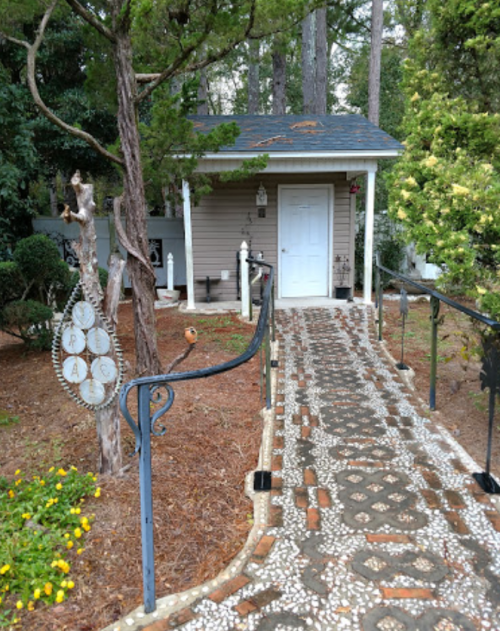 A charming pathway leads to a small, light-colored building surrounded by greenery and flowers.