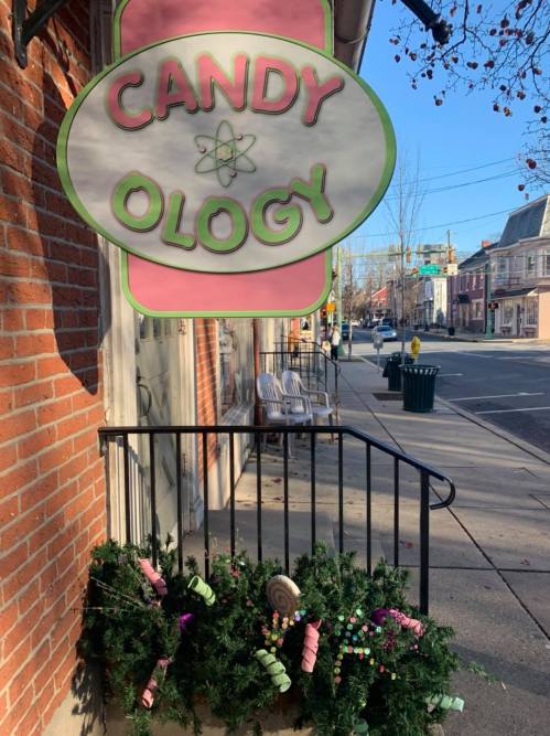 A colorful sign reading "CANDY OLOGY" hangs above a festive display of candy-themed decorations outside a shop.