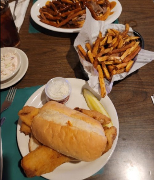 A plate with a fish sandwich, a side of coleslaw, and a pickle, alongside a basket of golden fries.