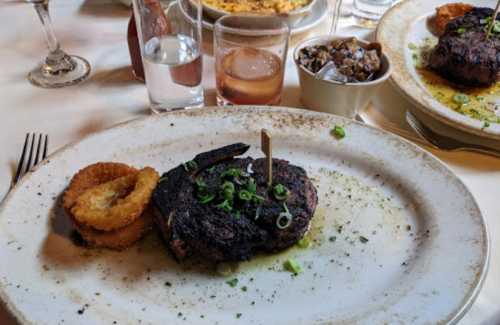 A plated steak with a toothpick, garnished with green onions, served with onion rings and a side of sauce.