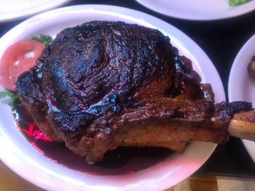 A large, well-cooked ribeye steak with a charred crust, garnished with a slice of tomato on a white plate.