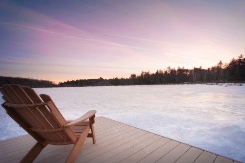 A wooden chair on a deck overlooking a snowy landscape at sunset, with soft pastel skies and trees in the distance.