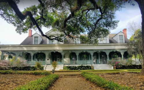 A large, historic house with a wraparound porch, surrounded by lush greenery and blooming flowers.