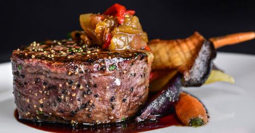 A perfectly cooked steak topped with sautéed vegetables and served with colorful roasted carrots on a white plate.