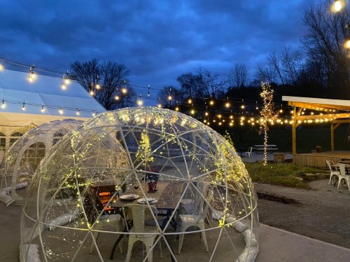 Clear domes with tables inside, illuminated by string lights, set against a twilight sky and a tent in the background.