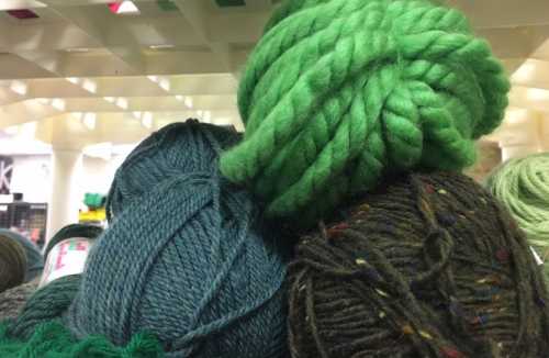 A close-up of various yarn balls in shades of green and blue, stacked together on a shelf.