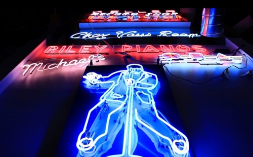 Colorful neon signs featuring "Riley Pianos," "Michael's," and a performer with a microphone against a dark background.