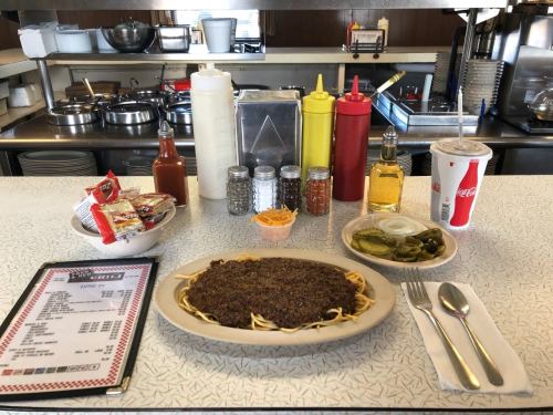 A plate of spaghetti topped with chili, surrounded by condiments, pickles, a drink, and a menu on a diner table.