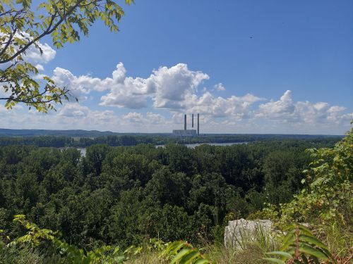 A power plant with three smokestacks is visible in the distance, surrounded by lush green trees and a blue sky with clouds.