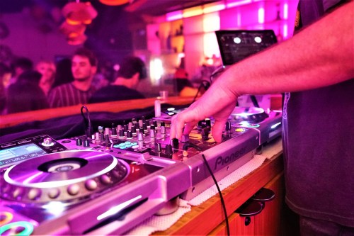 A DJ's hand adjusts the controls on a mixer, with colorful lights and a crowd in the background.