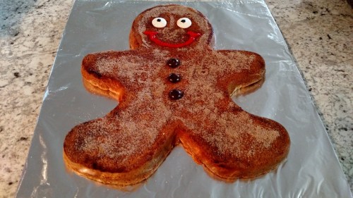 A large gingerbread man cookie with a smiling face, decorated with red icing and chocolate buttons, on a silver foil background.