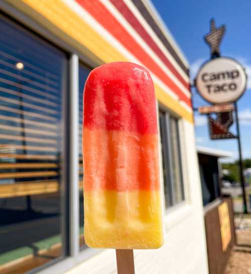 A colorful popsicle with red, orange, and yellow layers held up in front of a restaurant sign for Camp Taco.