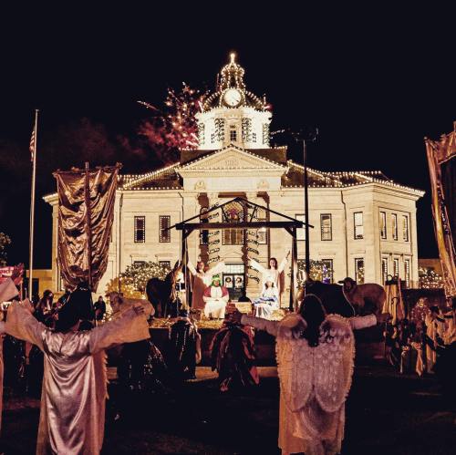 A festive scene at night with a decorated courthouse, performers in costumes, and fireworks in the background.