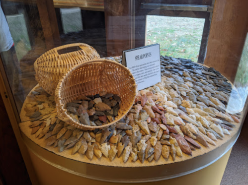 Display case featuring a woven basket and a variety of stone arrowheads arranged on a circular surface.