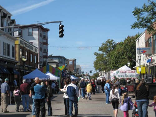 A busy street fair with vendors, people walking, and colorful tents under a clear blue sky.