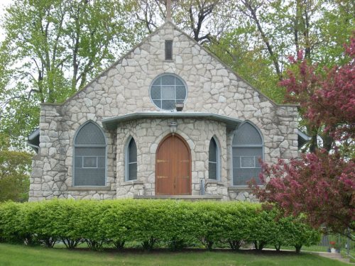 A stone church with arched windows and a wooden door, surrounded by green bushes and trees.