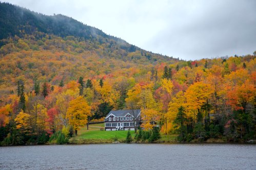 A scenic view of a house by a lake, surrounded by vibrant autumn foliage and mountains under a cloudy sky.