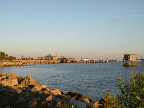 A calm waterfront scene with a pier, buildings in the background, and rocky shoreline under a clear sky.