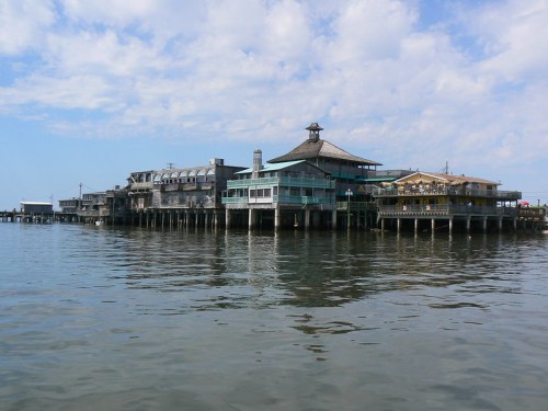 A waterfront building with a unique roof design, surrounded by calm water and a blue sky with scattered clouds.