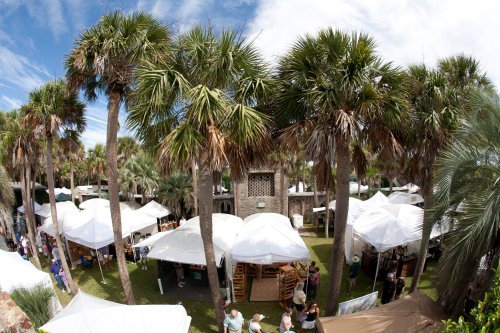 Aerial view of a vibrant outdoor market with white tents surrounded by palm trees and visitors exploring.