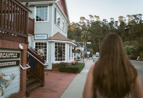 A woman walks along a sidewalk past a charming building with shops, surrounded by trees and a peaceful street scene.