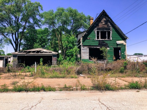 Abandoned green house with broken windows, overgrown vegetation, and a dilapidated garage under a clear blue sky.