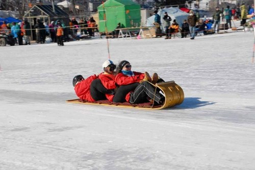 Three people in red jackets ride a wooden sled on ice, surrounded by spectators and tents in a winter setting.