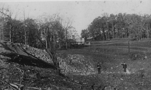 Black and white photo of a rocky landscape with two figures walking, surrounded by trees and a distant building.
