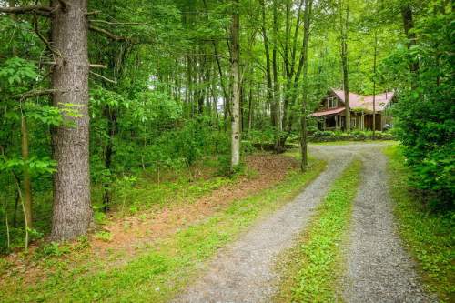 A gravel driveway leads to a cozy wooden house surrounded by lush green trees in a serene forest setting.