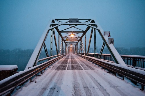 A snow-covered bridge at dusk, with streetlights illuminating the snowy path and a serene winter landscape.