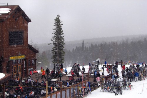 A snowy ski resort scene with people, ski equipment, and a wooden lodge in the background.