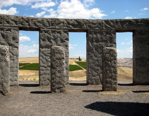 A stone structure with open frames overlooks a landscape of rolling hills and vineyards under a blue sky.