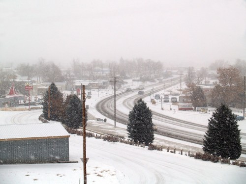 A snowy landscape with a winding road, trees, and buildings partially obscured by falling snow.