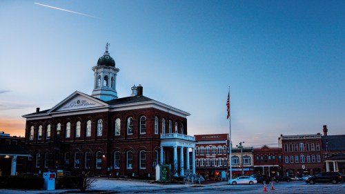 Historic brick building with a clock tower, American flag, and surrounding shops at dusk.