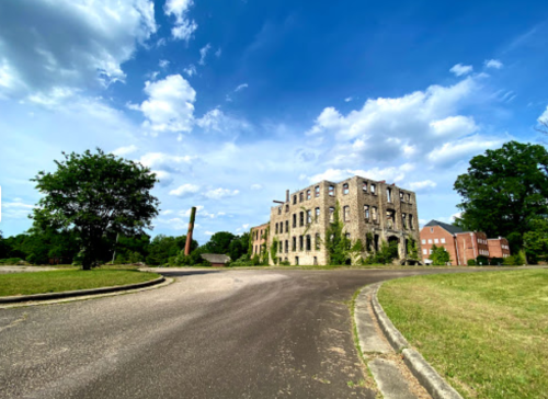 Abandoned building with a cloudy sky, surrounded by greenery and a winding road.