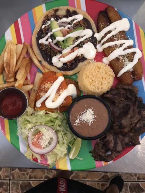 A colorful plate featuring tacos, rice, beans, fried plantains, salad, fries, and sauces.