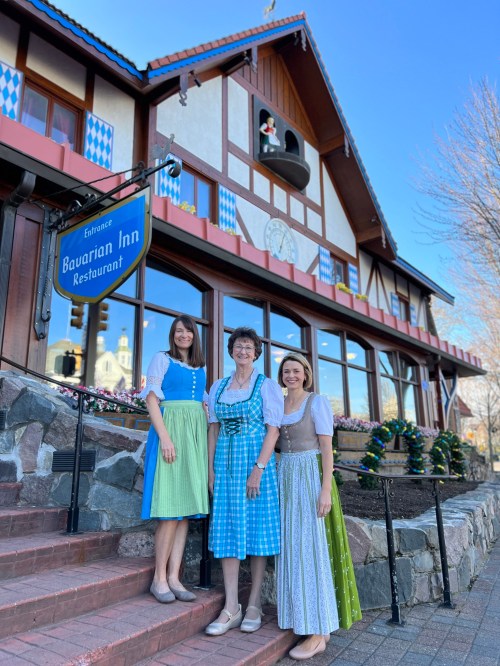 Three women in traditional Bavarian dresses stand in front of the Bavarian Inn Restaurant, with a blue sky above.