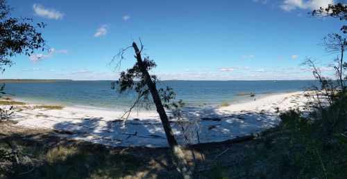 Panoramic view of a serene beach with white sand, calm blue water, and a clear sky dotted with clouds.