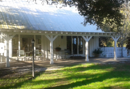 A charming white house with a glass roof, featuring a wooden deck and green chairs, surrounded by lush grass and trees.