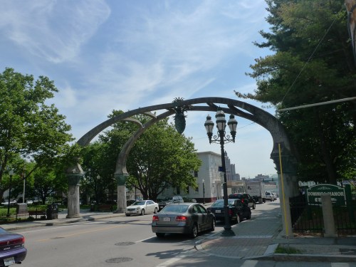 Archway with decorative elements and street lamps, surrounded by trees and buildings under a blue sky.